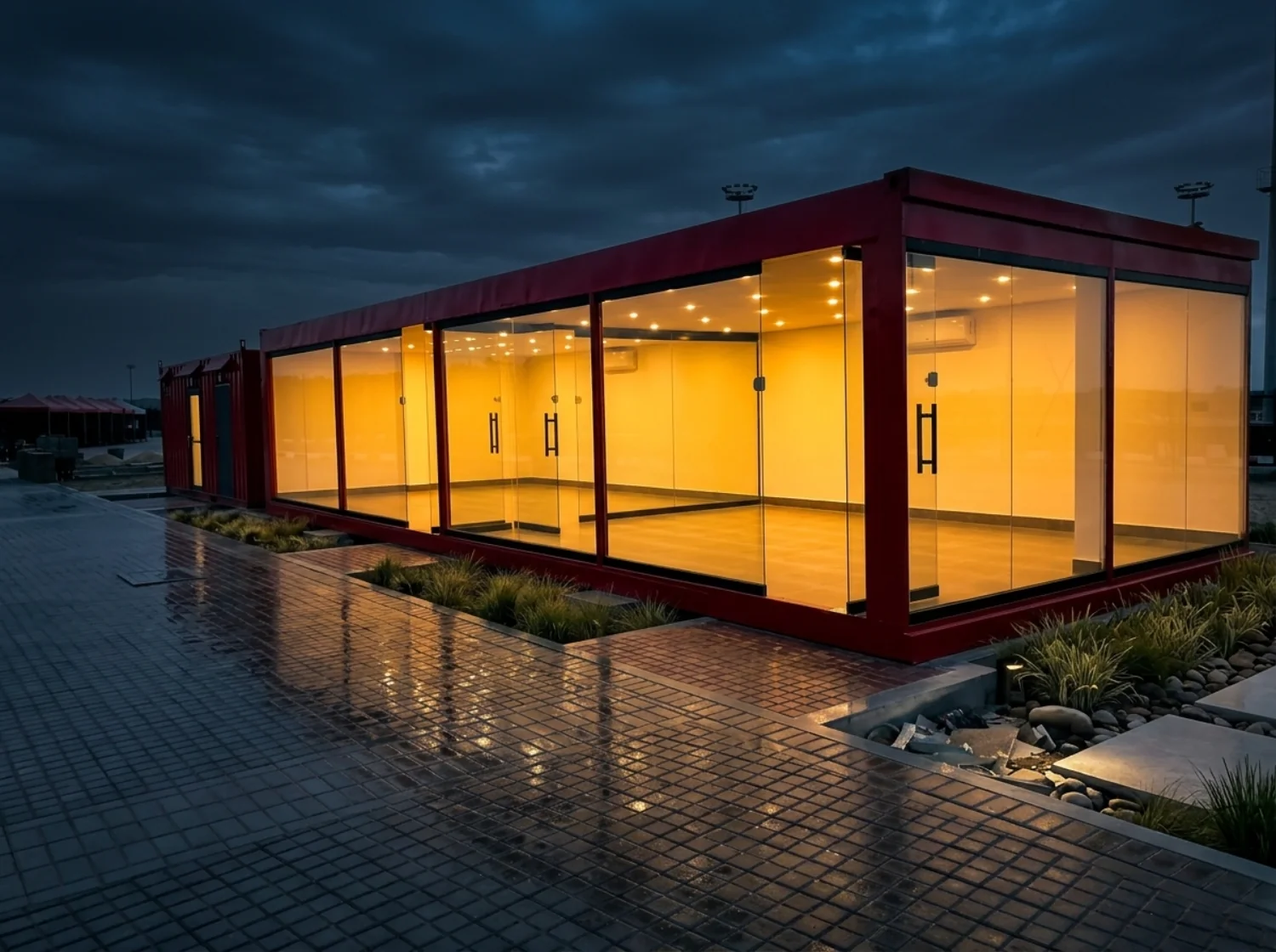 A red-framed container building with full-height glazing, lit warm inside, on wet paving at dusk.